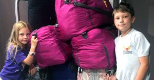Two kids standing against their luggage which is on a bell cart.